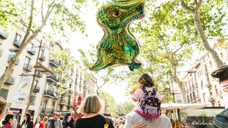A family, with their backs to the camera, strolling down La Rambla. The girl, sitting on her father's shoulders, is carrying a dragon-shaped balloon. 