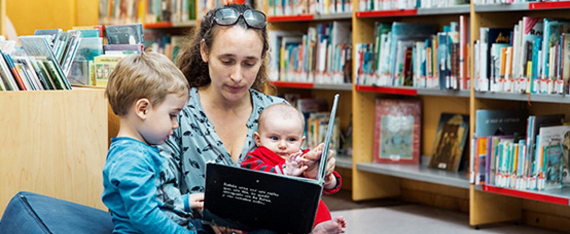 A woman, child and newborn baby sitting on cushions, looking at an illustrated album in a library 