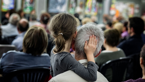 Una nena asseguda entre el públic d'una reunió veïnal posa la mà sobre el cap d'una persona anciana. 