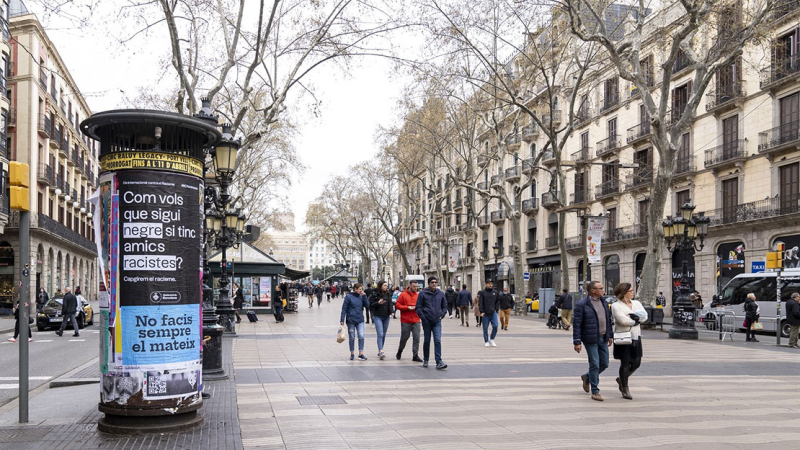 Totem en las Ramblas con el cartel de la campaña Barcelona antirracista que lleva el lema "¿Cómo quieres que sea negro si tengo amigos racistas?" y gente paseando por la calle