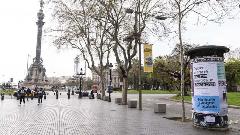 Totem al final de las Ramblas (Rambla de Santa Mónica), con el Monumento a Cristóbal Colón al fondo, con el cartel de Barcelona antirracista con el lema "Dejar entrar a los racistas provoca un efecto llamada"