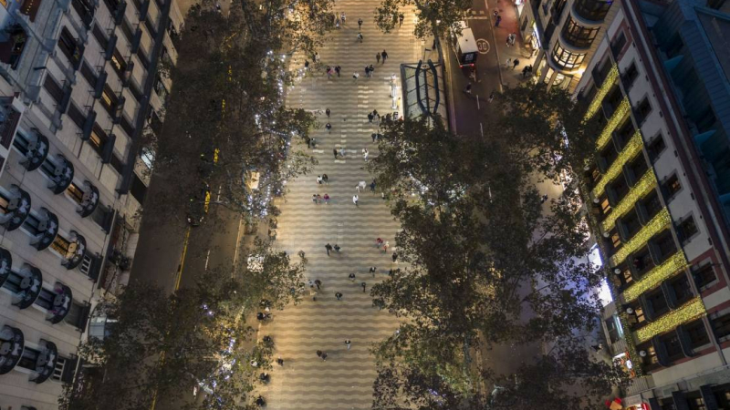 Vista aérea cenital de las Ramblas con gente paseando bajo los árboles iluminados con guirnaldas de luces de Navidad y entrando y saliendo de la estación de plaza de Catalunya