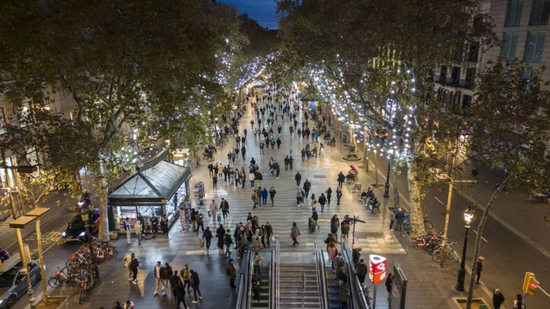 Vista aérea de las Ramblas con gente paseando bajo los árboles iluminados con guirnaldas de luces de Navidad y entrando y saliendo de la estación de plaza de Catalunya