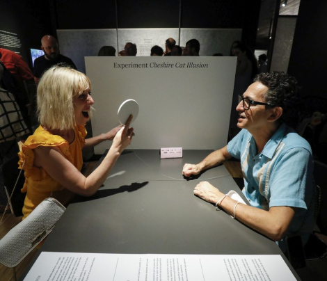 Dues persones assegudes en una taula una davant de l'altra experimenten una il·lusió òptica dins de l'exposició Cervell(s) al CCCB. © Martí E. Berenguer