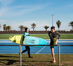 Una mujer camina con una tabla de surf y un hombre realiza estiramientos en la pista de atletismo del Centre Esportiu Municipal La Mar Bella