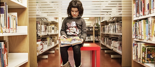 Nena asseguda sobre una taula llegint un llibre en un passadís de la biblioteca