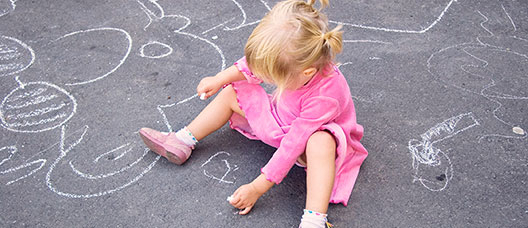 Nena asseguda al terra i fent dibuixos amb un guix al paviment 