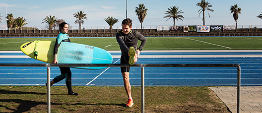 Una mujer camina con una tabla de surf y un hombre realiza estiramientos en la pista de atletismo del Centre Esportiu Municipal La Mar Bella