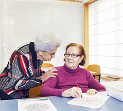 Dues usuàries d'un centre de dia parlant mentre una fa una activitat d'escriptura