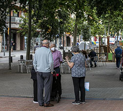 A group of elderly people talk standing on the Via Júlia. Around, you can see shops open and people sitting on the terraces or walking